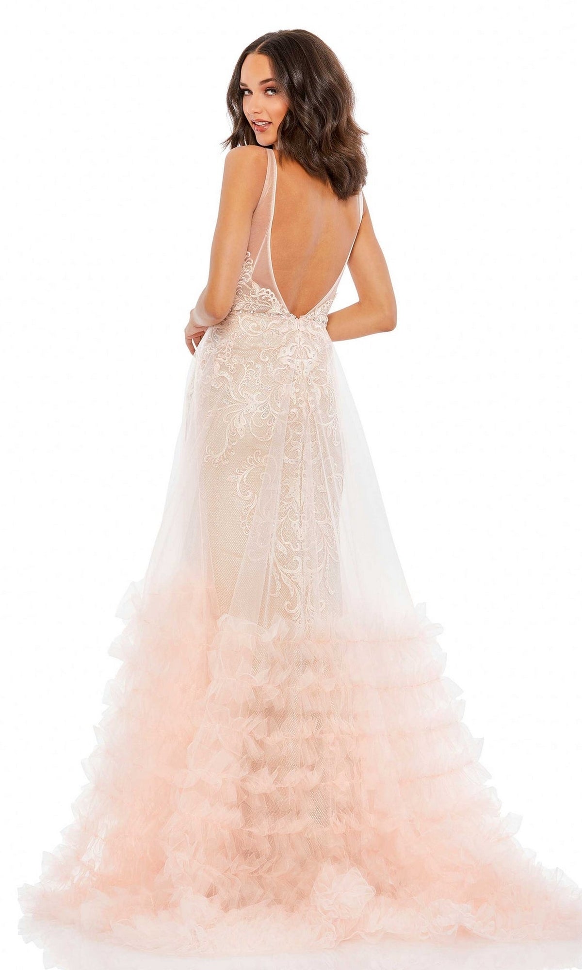 A woman stands facing slightly backward in the Long Formal Dress 11183 by Mac Duggal, a backless, sleeveless gown with a plunging v-neckline, lace detail, and layered tulle skirt in light pink and white.
